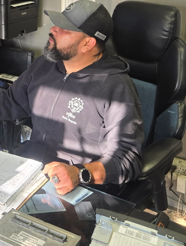A man at the desk in the shop office wearing an Alex’s Auto Repair hoodie, paperwork and an engine control unit on the desk in front of him.