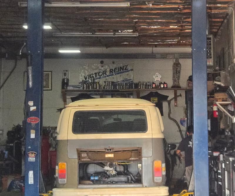A classic Volkswagen bus raised on the lift with its engine lid open, tool chests and a parts shelf along the wall.