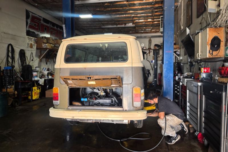 Inside the bay at 3596 Moline Street: a classic Volkswagen bus raised on a two-post lift with its rear engine lid open, and a technician crouched behind it working on the engine.