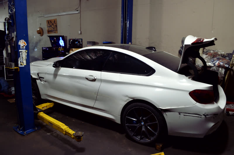 A white coupe raised on the two-post lift, seen from behind, its trunk lid open.