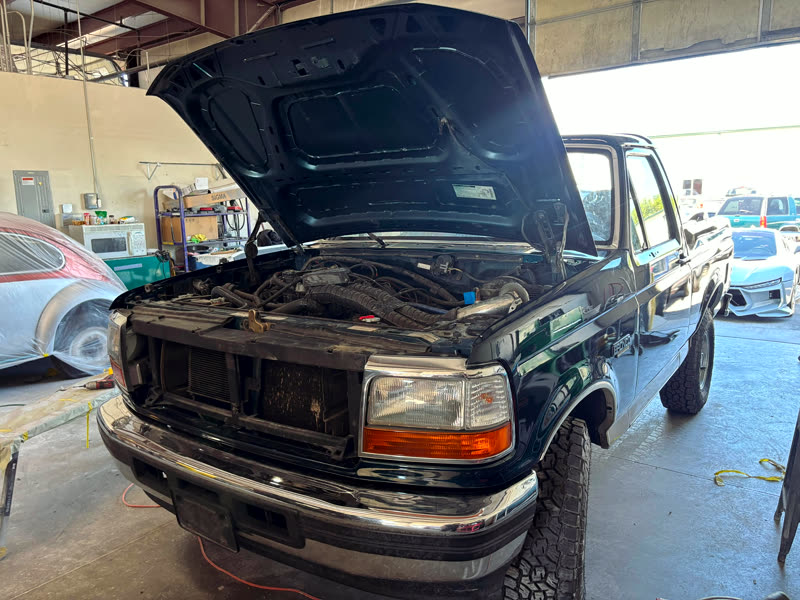An older Ford Bronco with its bonnet raised and its front trim removed, standing inside the shop.