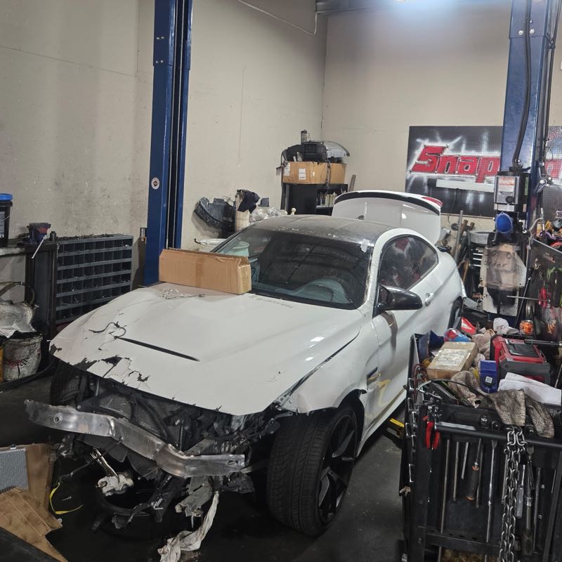 Inside the bay: a white coupe standing between the posts of a two-post lift, its bumper, hood and headlight assembly torn away by a front-end collision.