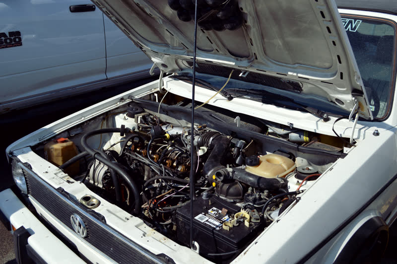 The engine compartment of an older Volkswagen, hoses and wiring across the bay, the badge visible on the front panel.