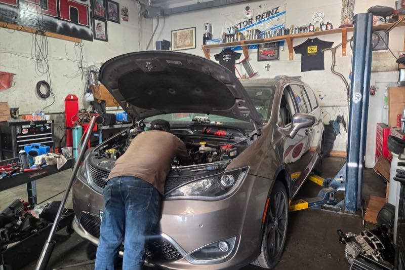 Inside the bay: a minivan up on a two-post lift with its hood raised, and a technician leaning into the engine bay with a tool in his hand.