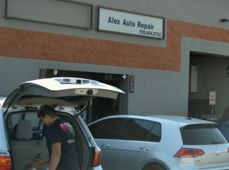 The front of the shop from the lot: the sign reading “Alex Auto Repair, 720.404.0732”, unit 103, two bay doors open, and a man working at the open tailgate of a car parked in front.