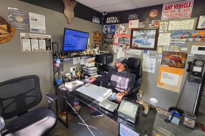 The front office at the shop: a desk with a monitor and paperwork, a wall of notices and dealer parts lists, and the open door through to the bay.
