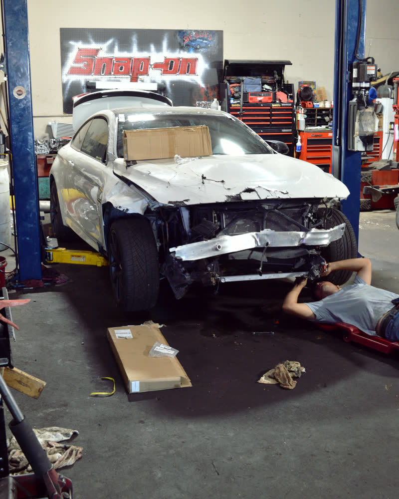 A white coupe with its whole front end stripped off, panels and bumper gone, a technician lying underneath it on a creeper.