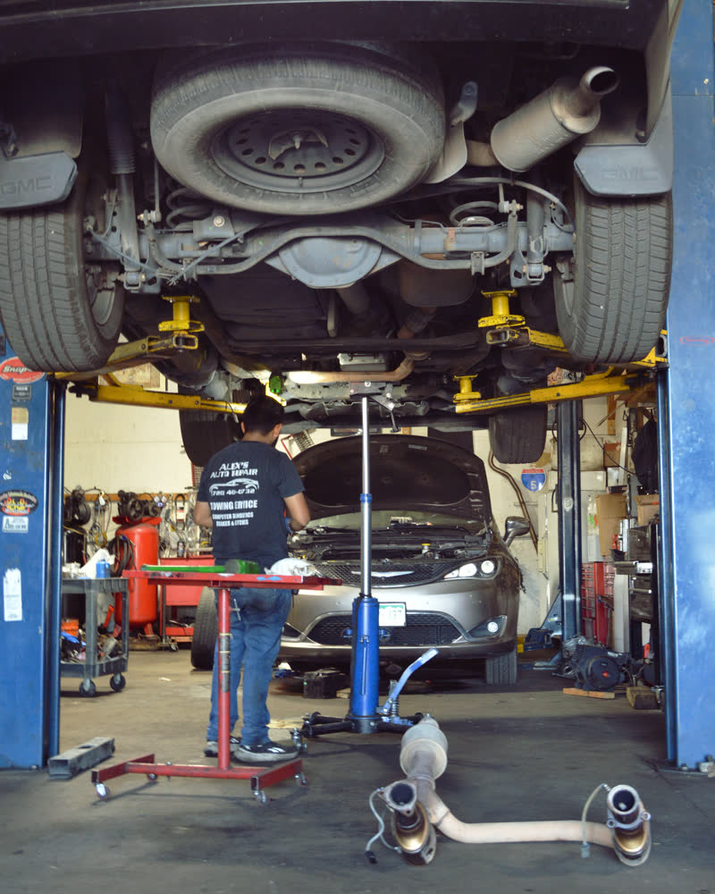 Looking up at a truck raised on the lift, exhaust and driveshaft overhead, a technician below with a transmission jack.