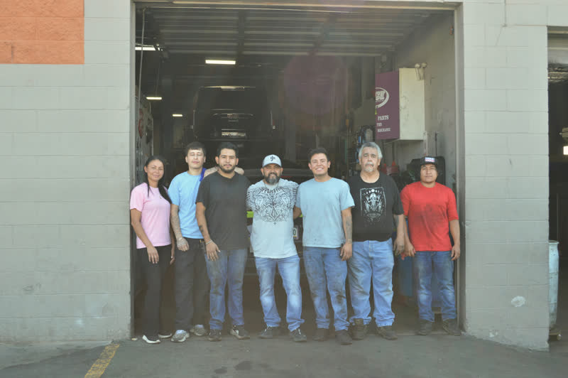 Seven people standing across the open bay door of the shop, a truck up on the lift behind them.